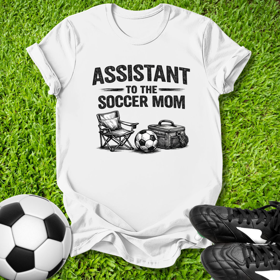 White t-shirt with 'Assistant to the Soccer Mom' graphic on grass with soccer ball and cleats.