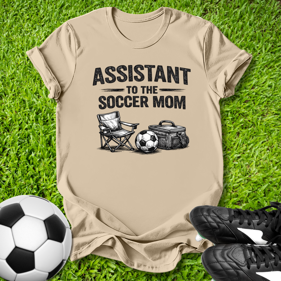Beige t-shirt with 'Assistant to the Soccer Mom' text and graphics on grass with soccer ball and shoes.