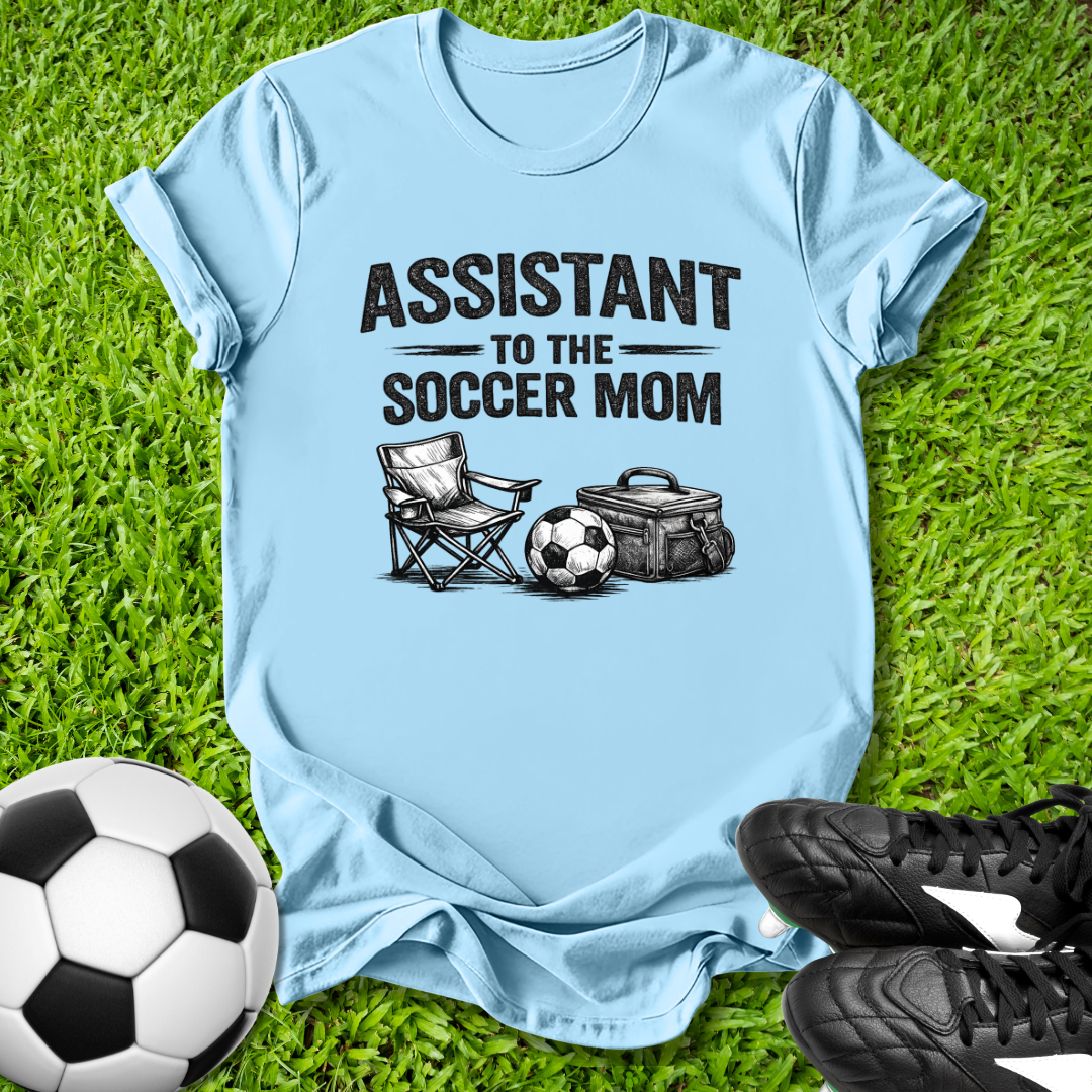 Light blue t-shirt with 'Assistant to the Soccer Mom' text and graphics on grass with soccer ball and shoes.