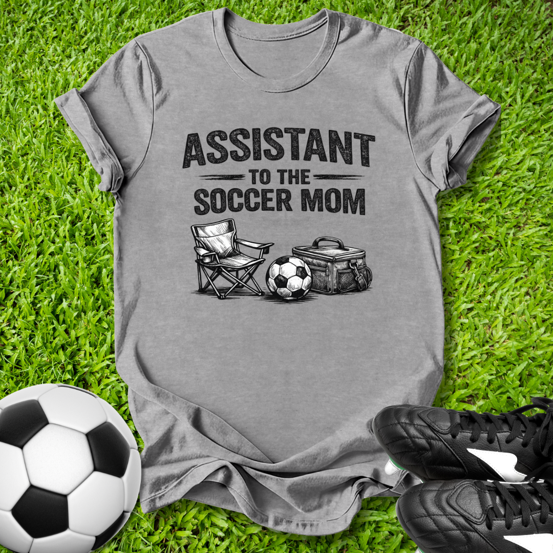 Gray t-shirt with 'Assistant to the Soccer Mom' text on grass with soccer ball and cleats.