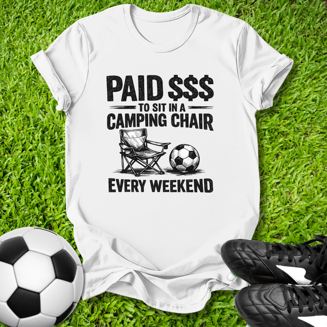 White t-shirt with a graphic and text on grass with soccer balls and shoes.
