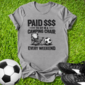 Gray t-shirt with a graphic and text on grass with soccer balls and shoes.