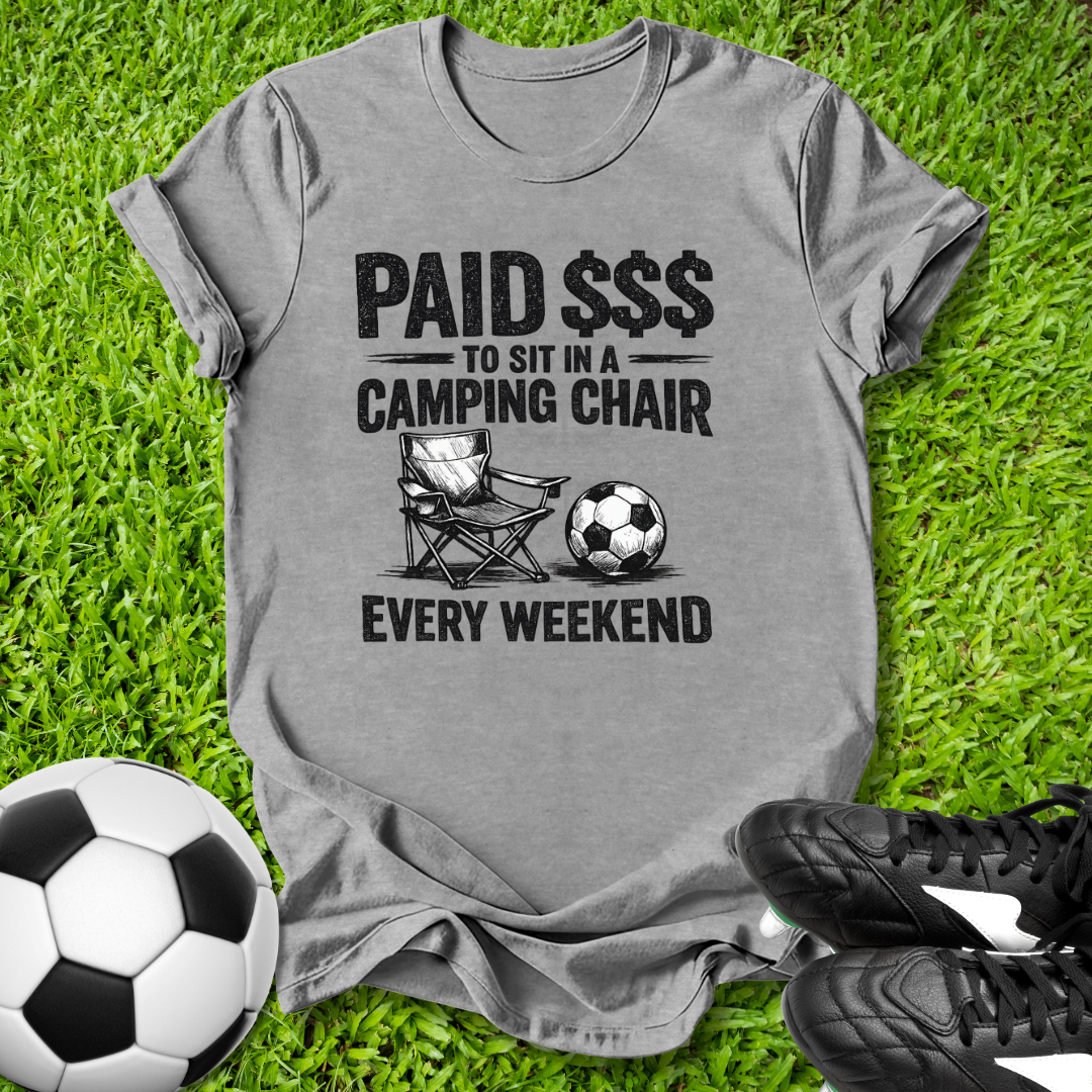 Gray t-shirt with a graphic and text on grass with soccer balls and shoes.