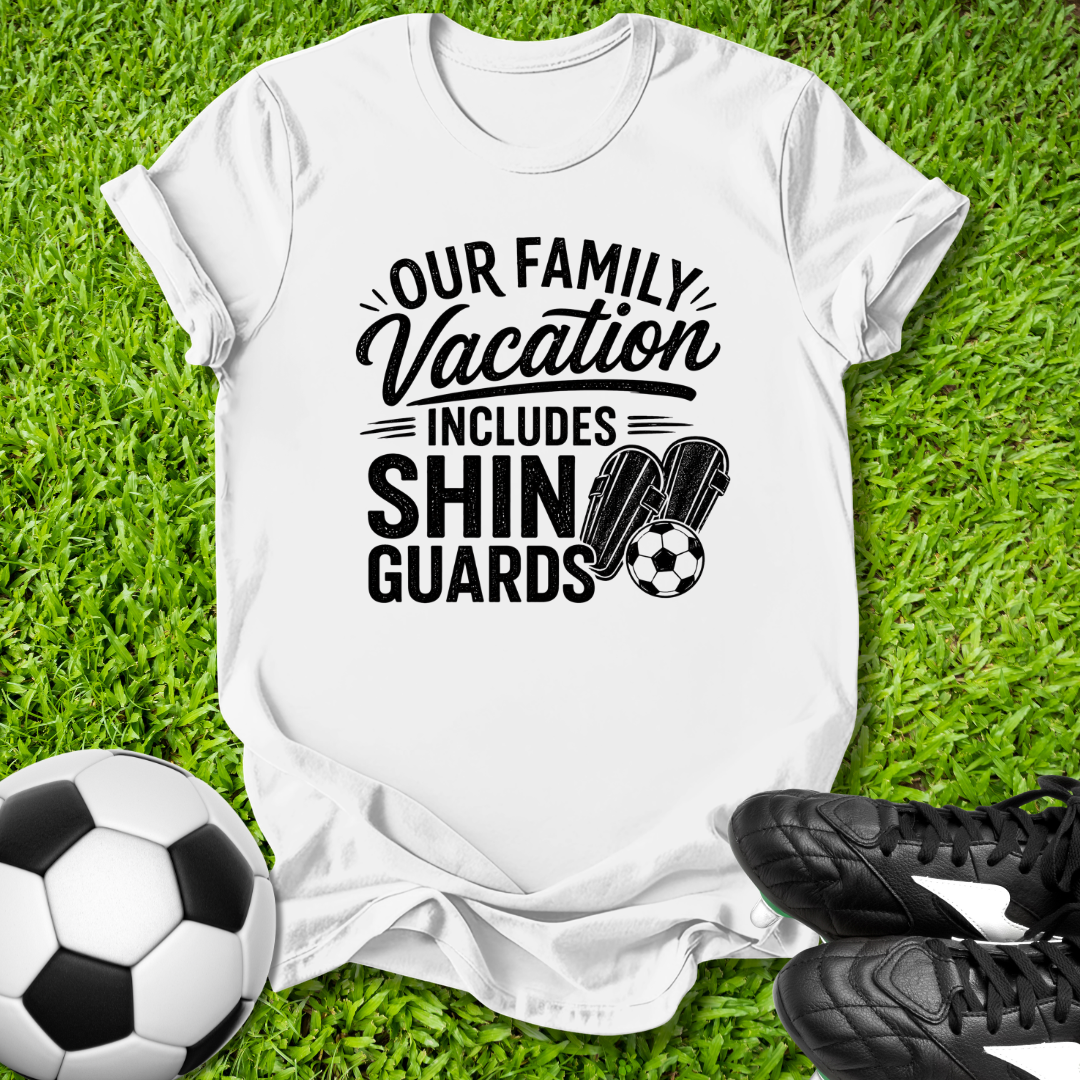 White t-shirt with 'Our Family Vacation Includes Shin Guards' text on grass with soccer ball and shoes.