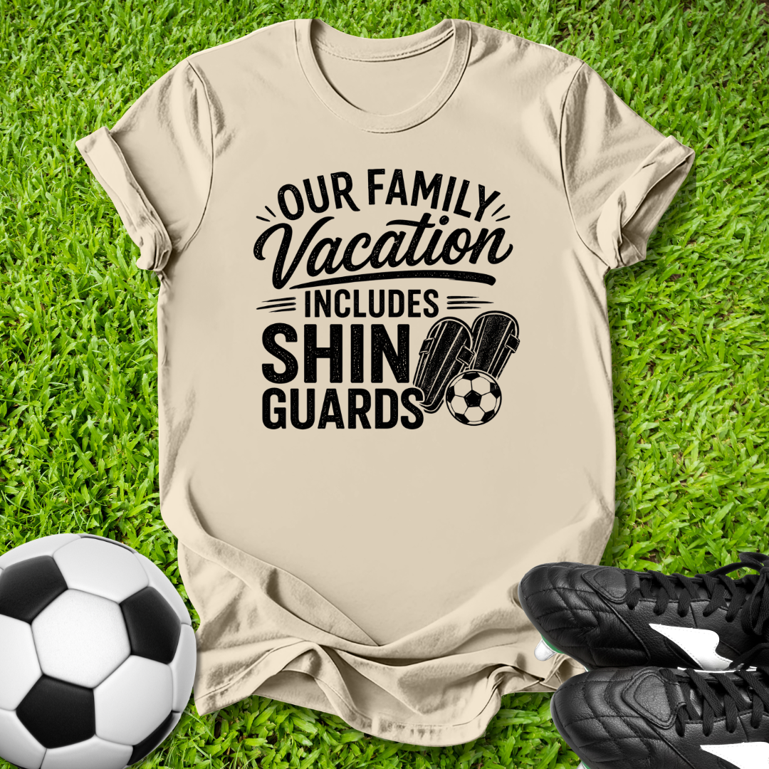 Beige t-shirt with 'Our Family Vacation Includes Shin Guards' text on grass with soccer ball and shoes.
