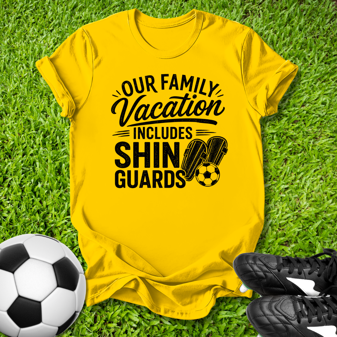 Yellow t-shirt with 'Our Family Vacation Includes Shin Guards' text on grass with soccer ball and shoes.