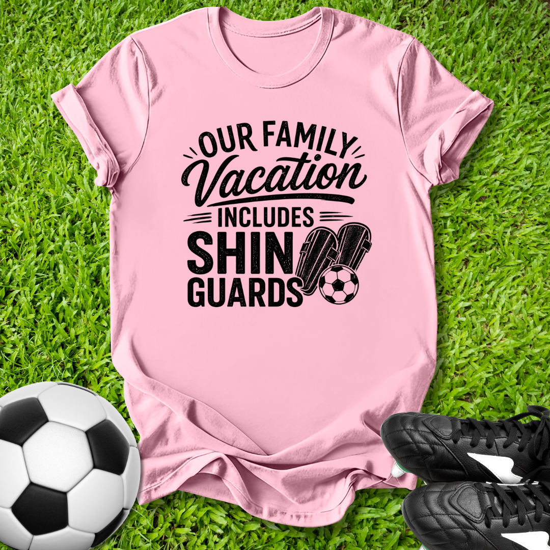 Pink t-shirt with 'Our Family Vacation Includes Shin Guards' text on grass with soccer ball and shoes.