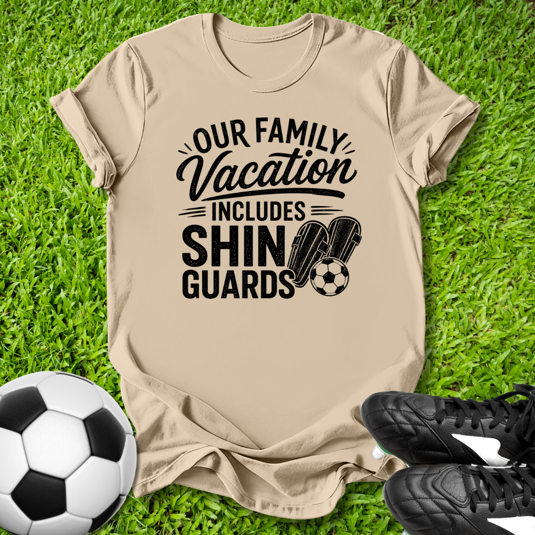 Beige t-shirt with 'Our Family Vacation Includes Shin Guards' text on grass with soccer ball and shoes.