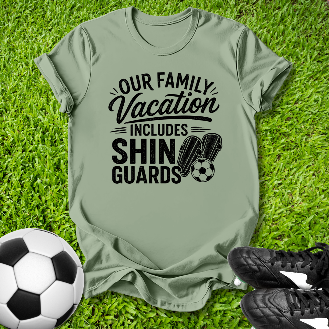 Green t-shirt with 'Our Family Vacation Includes Shin Guards' text on grass with soccer ball and shoes.