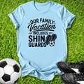 Light blue t-shirt with 'Our Family Vacation Includes Shin Guards' text on grass with soccer ball and shoes.