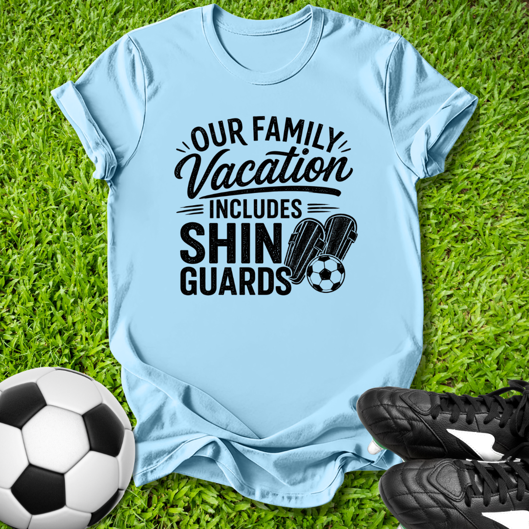 Light blue t-shirt with 'Our Family Vacation Includes Shin Guards' text on grass with soccer ball and shoes.