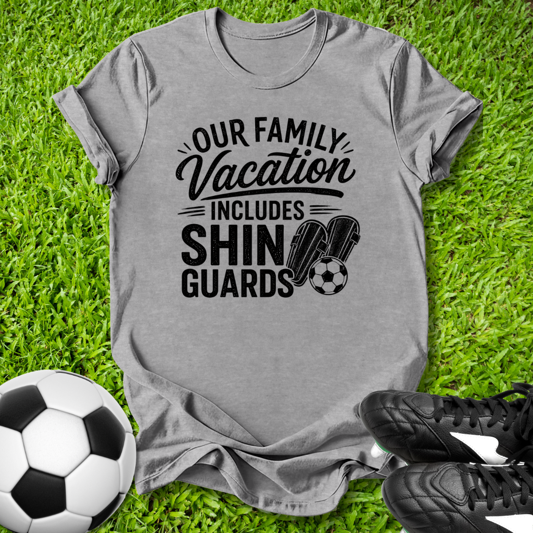Gray t-shirt with 'Our Family Vacation Includes Shin Guards' text on grass with soccer ball and shoes.