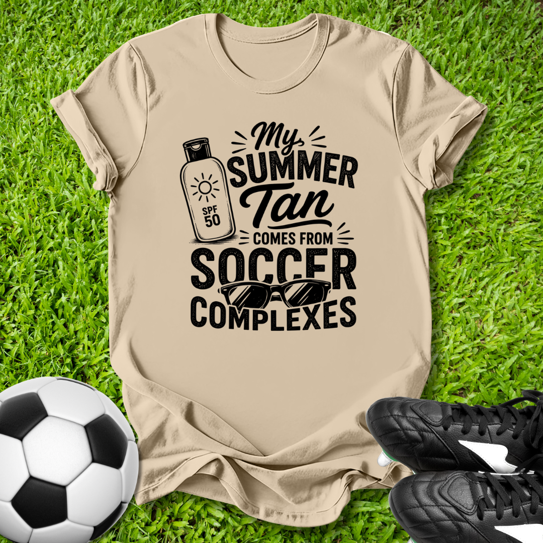 Beige t-shirt with 'My Summer Tan Comes from Soccer Complexes' text on grass with soccer ball and shoes.