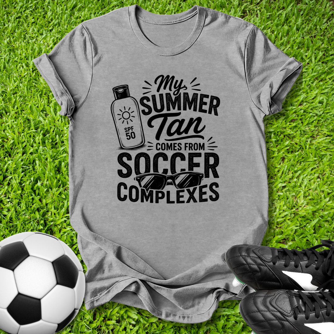 Gray t-shirt with 'My Summer Tan Comes from Soccer Complexes' text on grass with soccer ball and shoes.