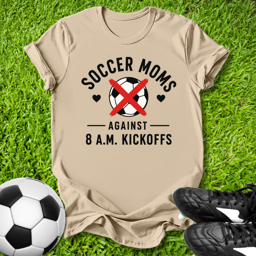 Beige t-shirt with 'Soccer Moms Against 8 A.M. Kickoffs' design on grass with soccer ball and shoes.