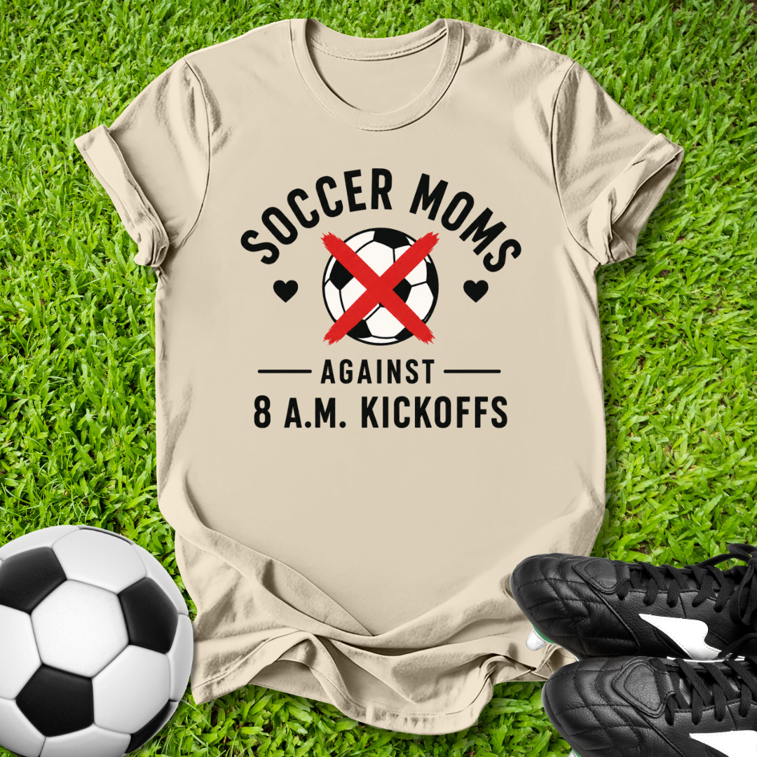 Beige tt-shirt with 'Soccer Moms Against 8 A.M. Kickoffs' design on grass with soccer ball and shoes.