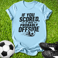 Light blue t-shirt with soccer-themed text on grass with soccer ball and shoes.