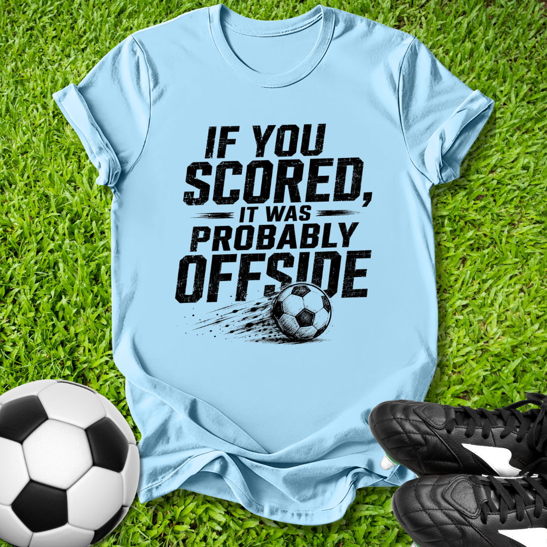 Light blue t-shirt with soccer-themed text on grass with soccer ball and shoes.