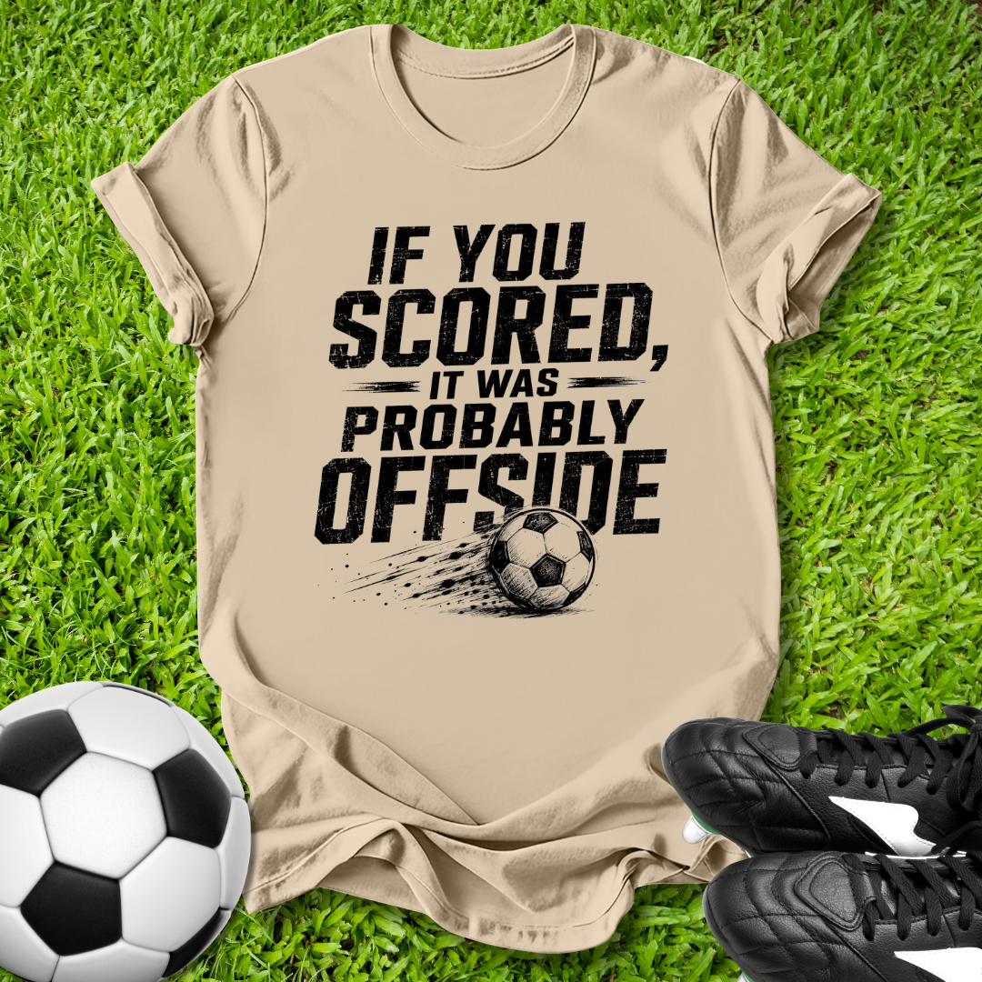 Beige t-shirt with offside soccer-themed text on grass with soccer ball and shoes.
