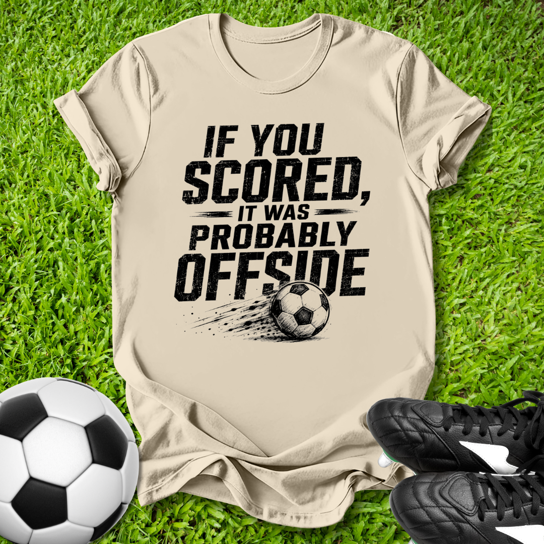 Beige t-shirt with offside soccer-themed text on grass with soccer balls and shoes.