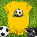 Yellow t-shirt with soccer ball graphic on grass, next to a real soccer ball and black shoes.