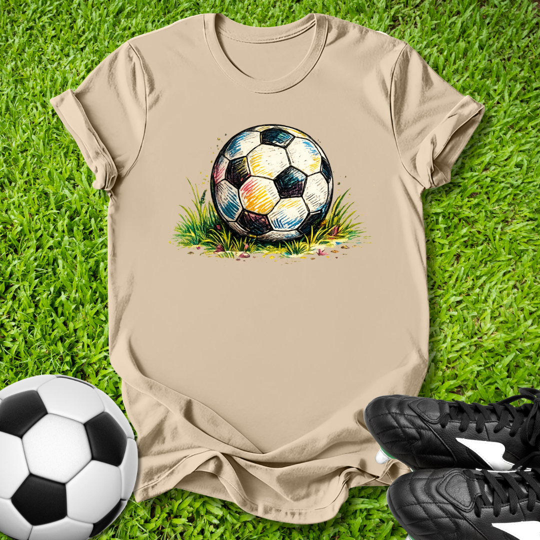 Beige t-shirt with a colorful soccer ball graphic on grass, next to a real soccer ball and black cleats.