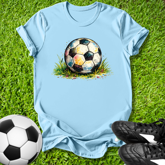 Light blue t-shirt with a colorful soccer ball graphic on grass, next to a real soccer ball and cleats.