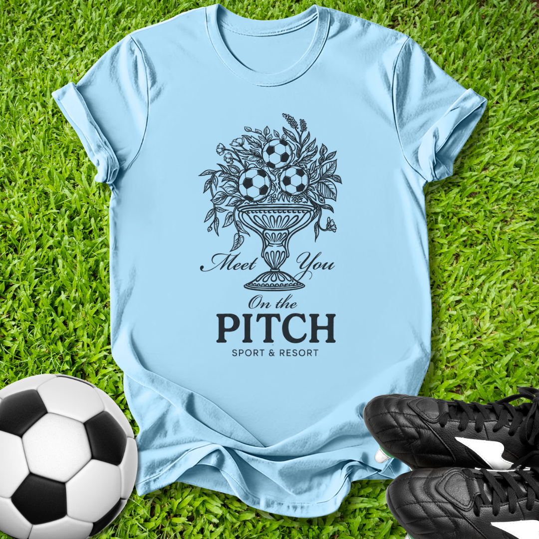 Light blue t-shirt with sports-themed design on grass with soccer ball and shoes.