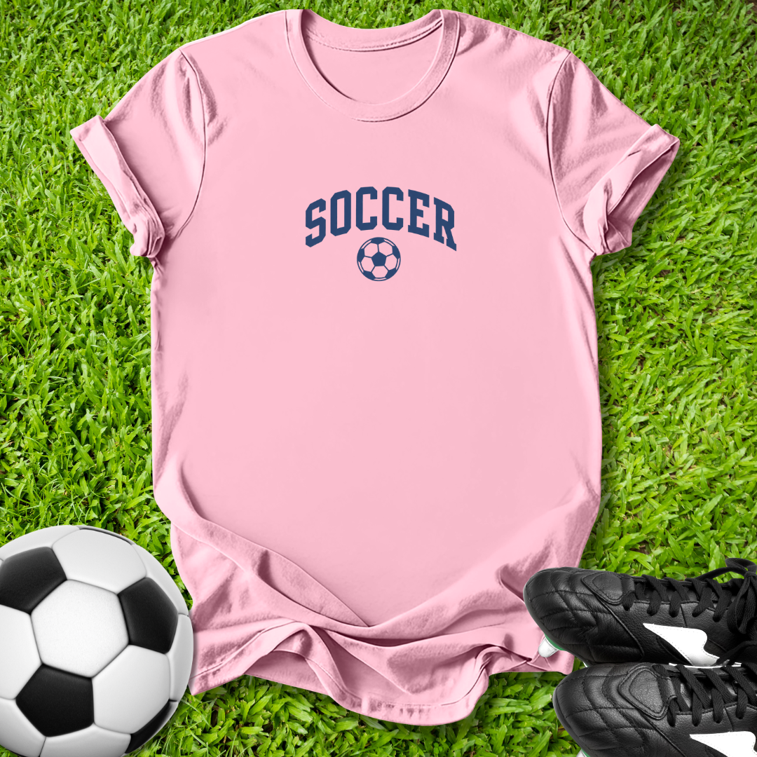 Pink t-shirt with 'SOCCER' text on grass with soccer ball and shoes