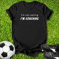 Black t-shirt with 'I'm not yelling I'm coaching' text on grass with soccer ball and shoes.