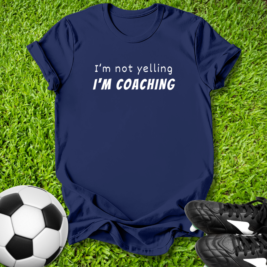 Navy blue t-shirt with 'I'm not yelling I'm coaching' text on grass with soccer ball and shoes.