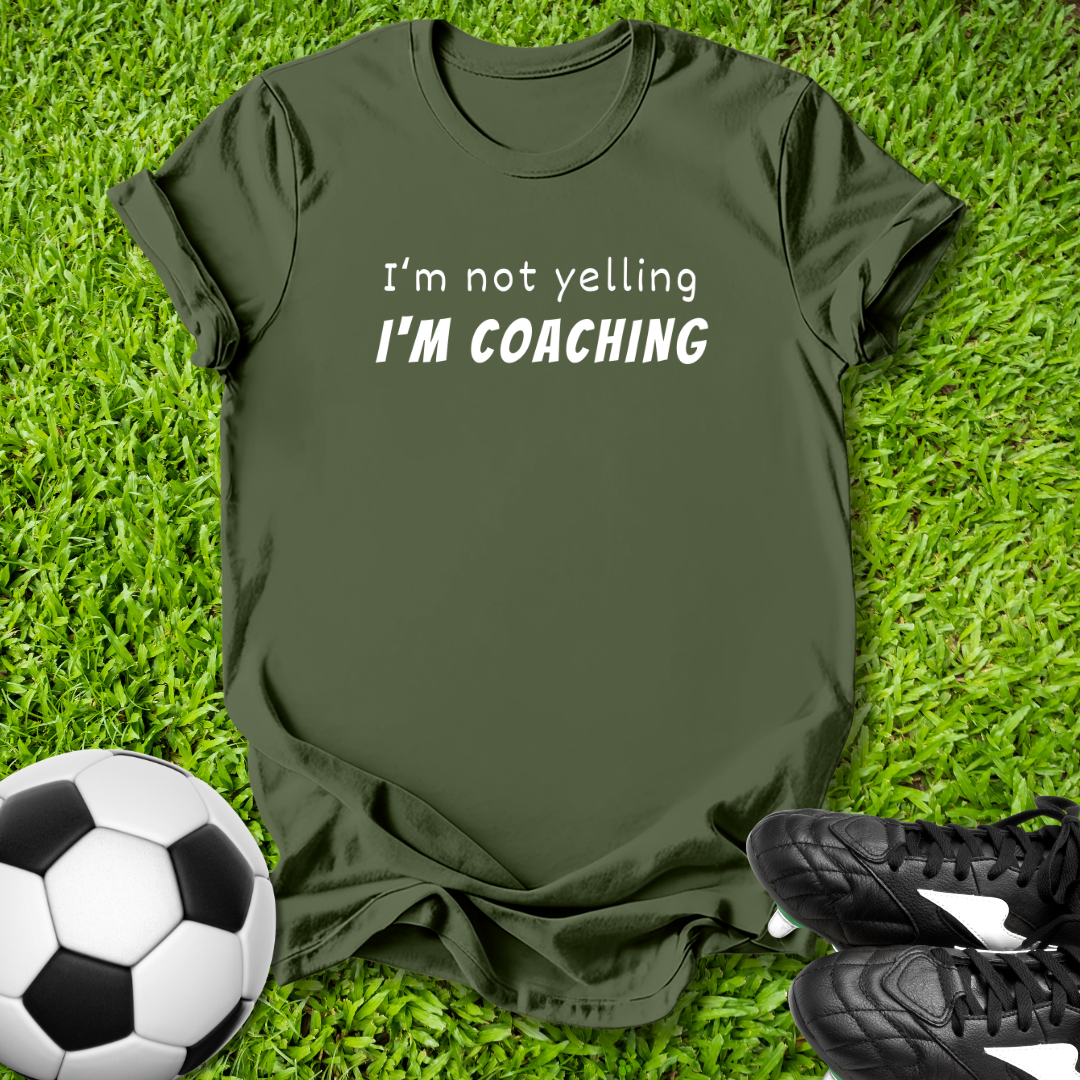 Green t-shirt with 'I'm not yelling I'm coaching' text on grass with soccer ball and shoes.