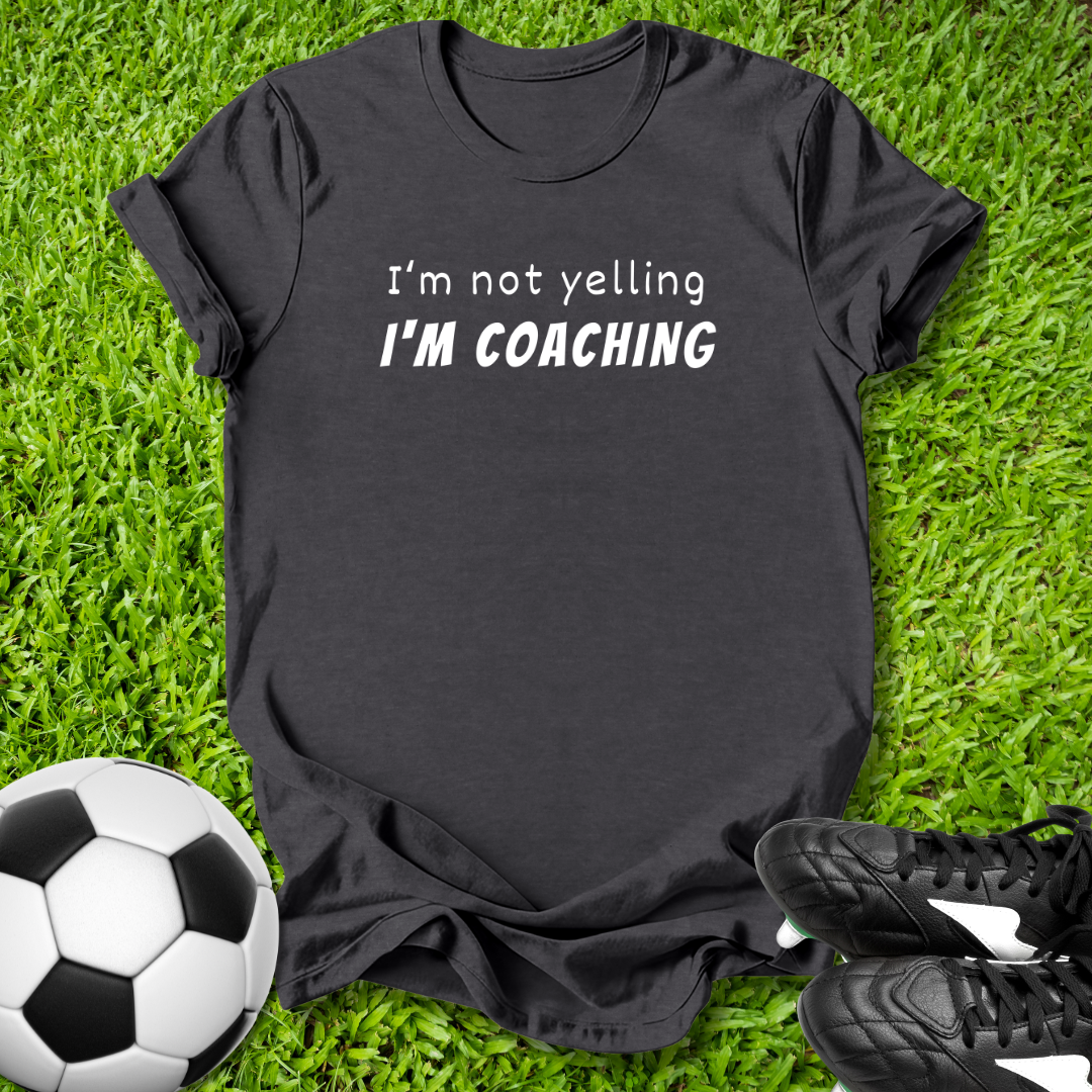 Black t-shirt with 'I'm not yelling I'm coaching' text on grass with soccer ball and shoes.