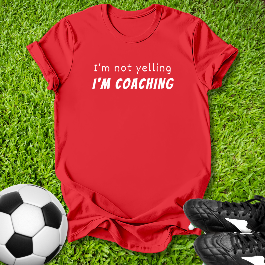 Red t-shirt with 'I'm not yelling I'm coaching' text on grass with soccer ball and shoes.