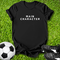 Black t-shirt with 'MAIN CHARACTER' text on grass with soccer ball and shoes