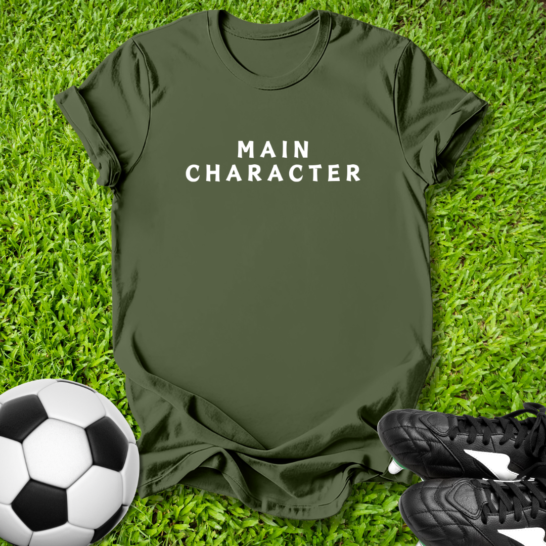 Green t-shirt with 'MAIN CHARACTER' text on grass with soccer ball and shoes