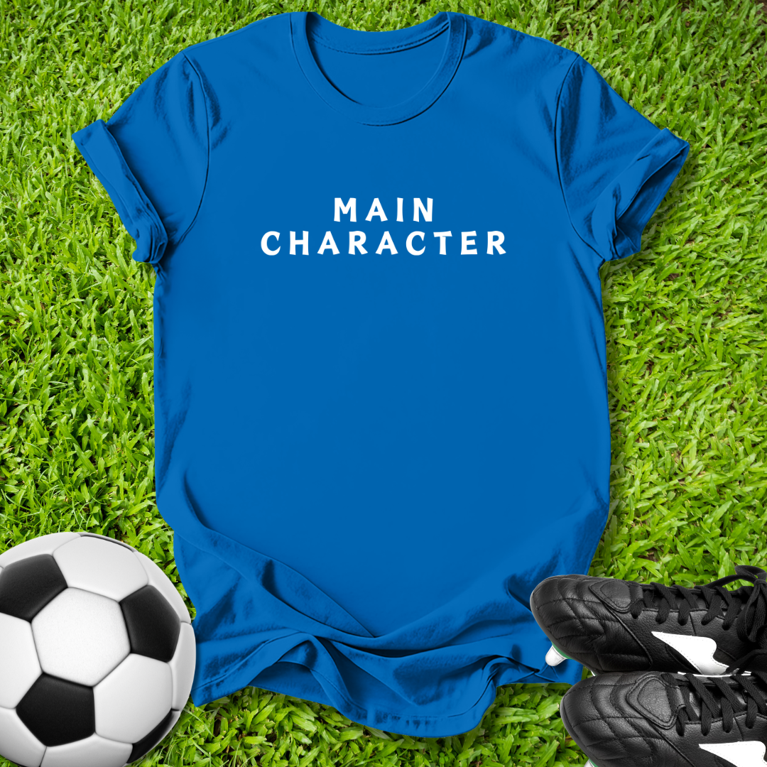 Blue t-shirt with 'MAIN CHARACTER' text on grass with soccer ball and shoes