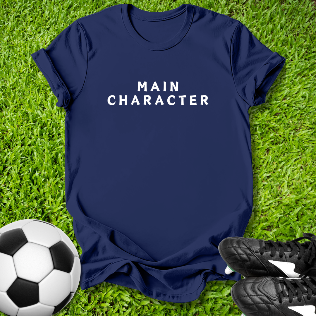 Navy blue t-shirt with 'MAIN CHARACTER' text on grass with soccer ball and shoes