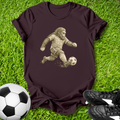 Maroon t-shirt with a graphic of a gorilla playing soccer on grass with a soccer ball and shoes.
