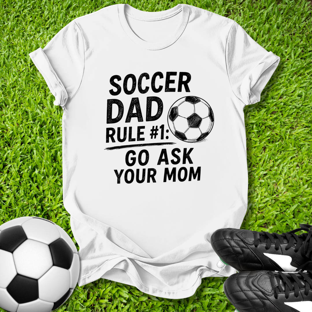White t-shirt with 'Soccer Dad Rule #1: Go Ask Your Mom' text on grass with soccer balls and shoes.