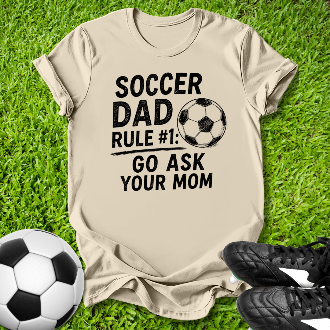 Beige t-shirt with 'Soccer Dad' text on grass with soccer ball and shoes