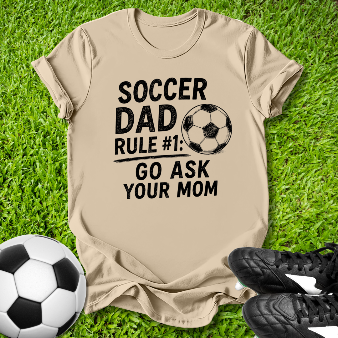 Beige t-shirt with 'Soccer Dad' text on grass with soccer ball and shoes.