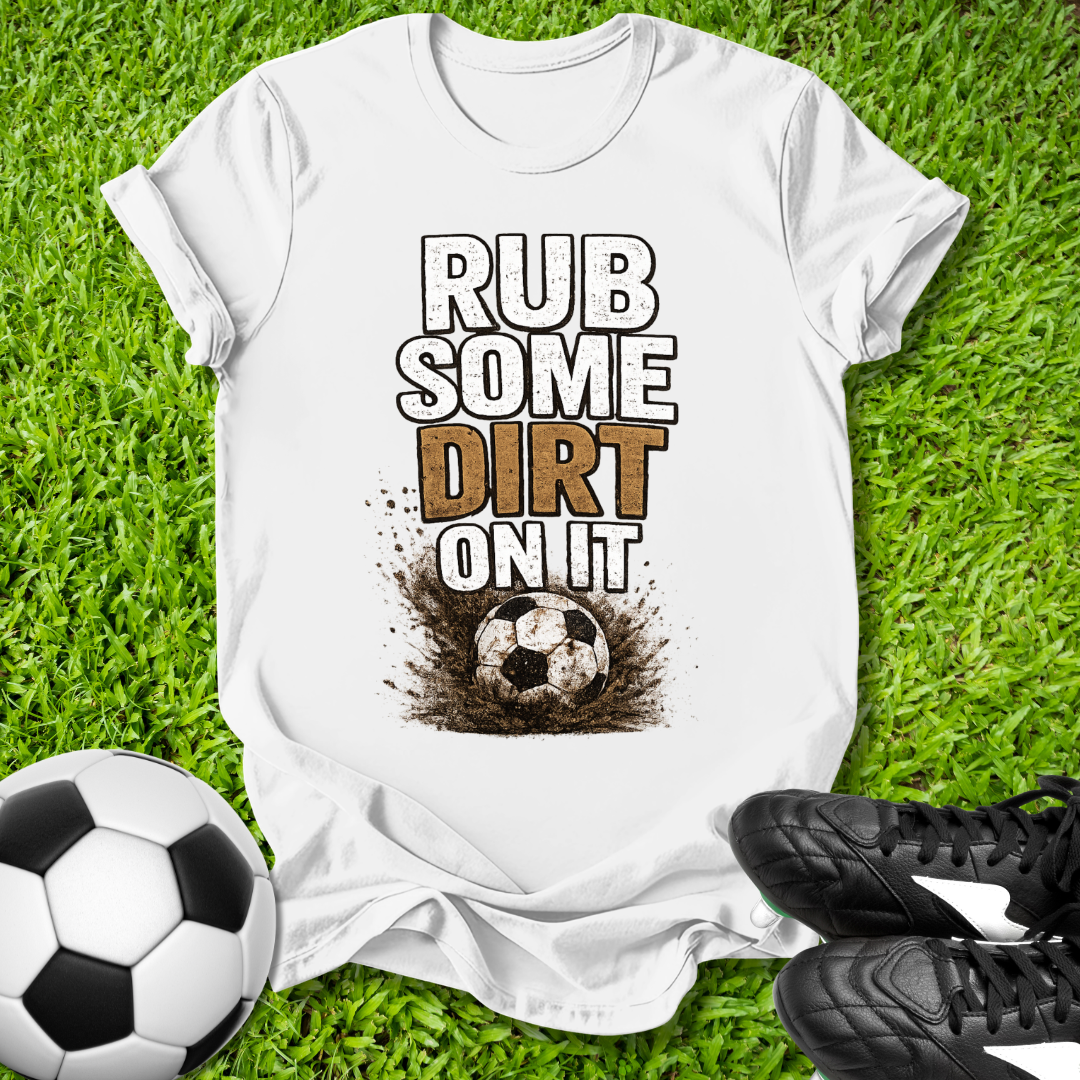 White t-shirt with 'Rub some dirt on it' text on grass with soccer ball and shoes.