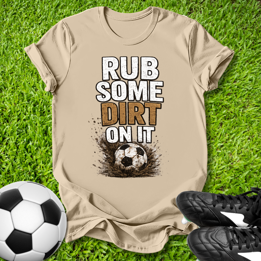 Beige  t-shirt with 'Rub some dirt on it' text on grass with soccer ball and shoes.