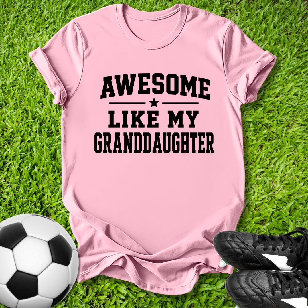 Pink t-shirt with 'Awesome like my granddaughter' text on grass with soccer ball and shoes.
