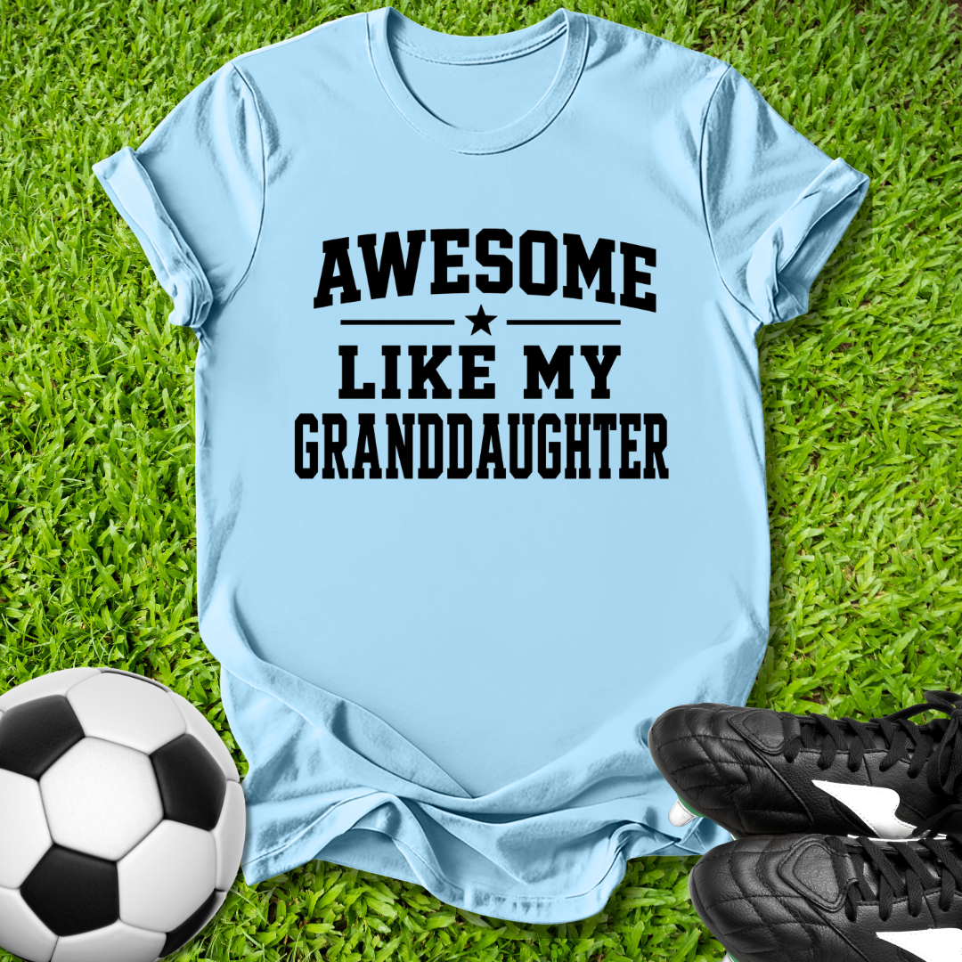 Light blue t-shirt with 'Awesome like my granddaughter' text on grass with soccer ball and shoes.