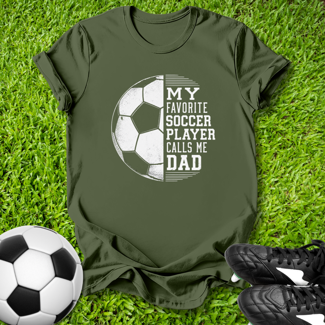 Green t-shirt with soccer ball graphic and text on grass with soccer ball and shoes