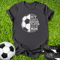 Black t-shirt with soccer ball graphic and text on grass with soccer ball and shoes