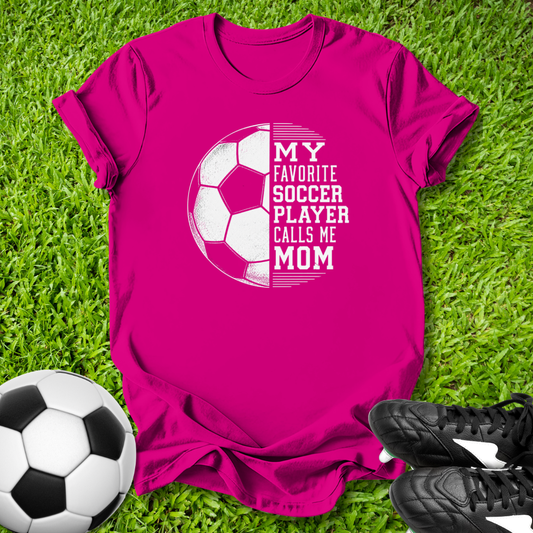 Pink t-shirt with soccer graphic and text on grass with soccer ball and shoes