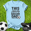 Light blue t-shirt with 'This is what an amazing soccer dad looks like' text on grass with soccer ball and shoes.